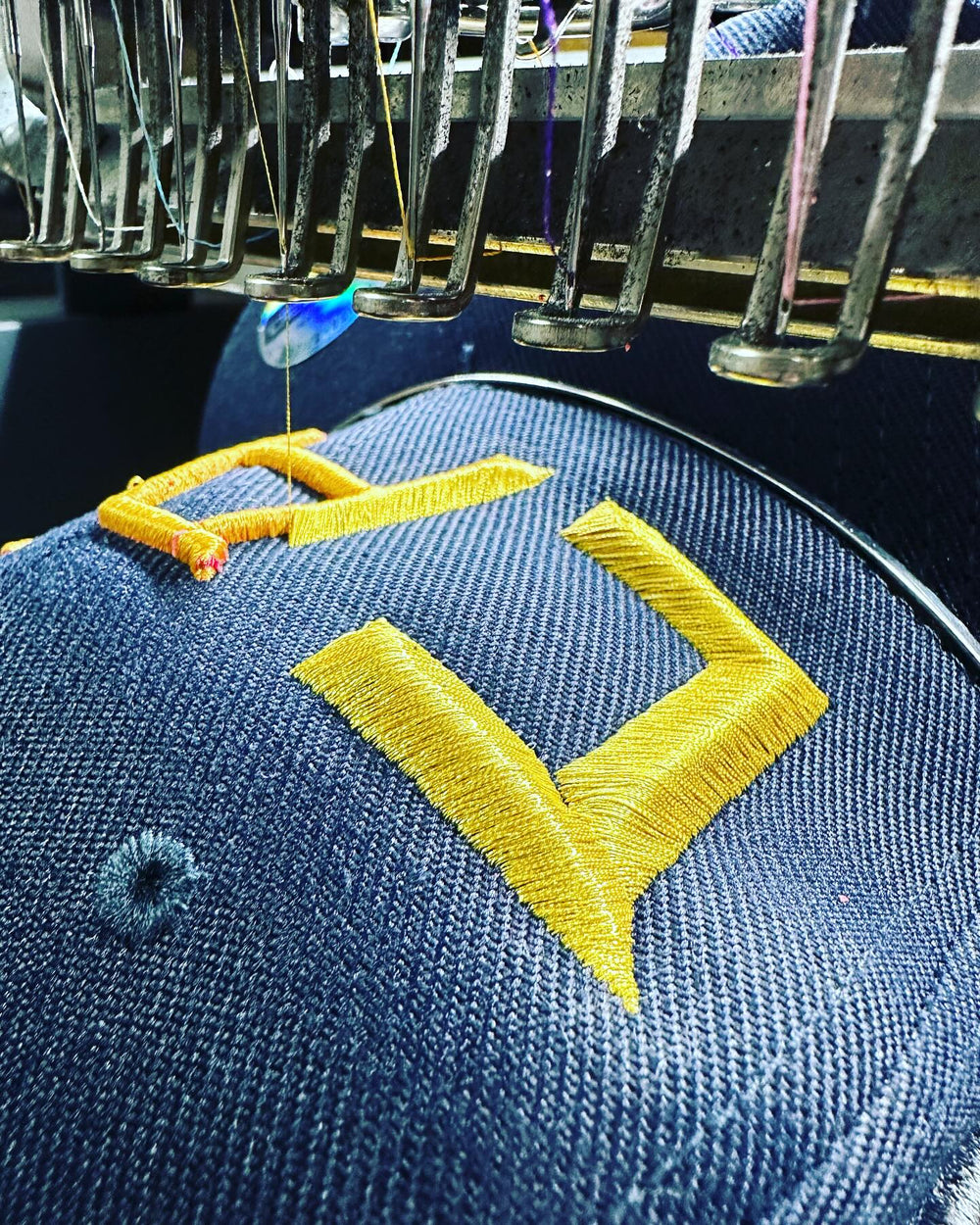 Embroidery hoop with blue fabric and yellow letter 'V' design, surrounded by sewing machine needles.