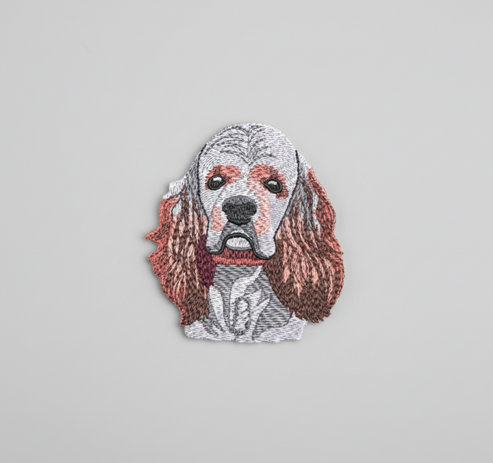 Realistic American Cocker Spaniel embroidery patch featuring a detailed dog face with long floppy ears in white, brown, and rust tones, stitched with textured thread shading on a light background, original EmbroMania artwork.