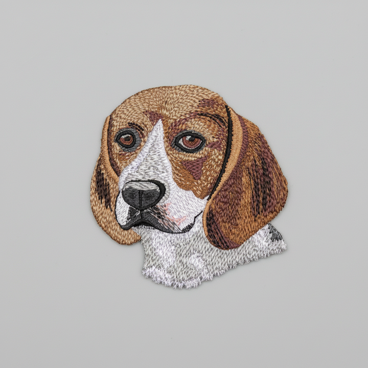 Realistic beagle embroidery patch featuring a detailed dog portrait with brown and white coat, floppy ears, and expressive eyes, stitched with textured thread shading on a light background, original EmbroMania artwork.