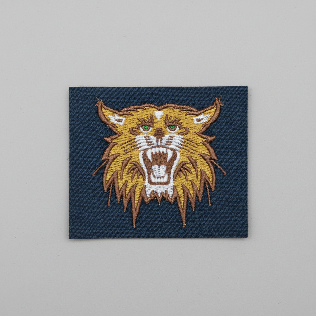 Bobcat embroidery patch featuring a fierce wildcat head with open mouth, sharp fangs, and green eyes in detailed brown, gold, and white stitching on a dark fabric background, original EmbroMania artwork.