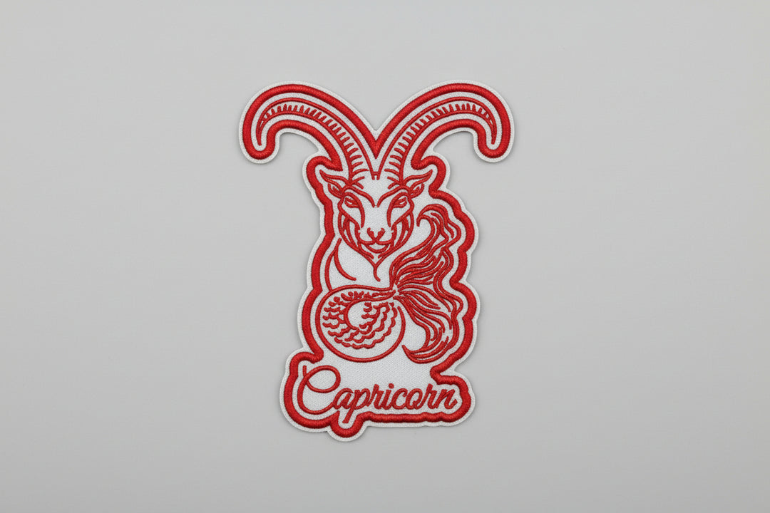 Capricorn zodiac sea-goat embroidery patch featuring a stylized goat with long curved horns and a curled tail in red outline stitching with the word “Capricorn” in script below, on a light background, original EmbroMania artwork.