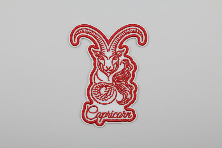 Capricorn zodiac sea-goat embroidery patch featuring a stylized goat with long curved horns and a curled tail in red outline stitching with the word “Capricorn” in script below, on a light background, original EmbroMania artwork.
