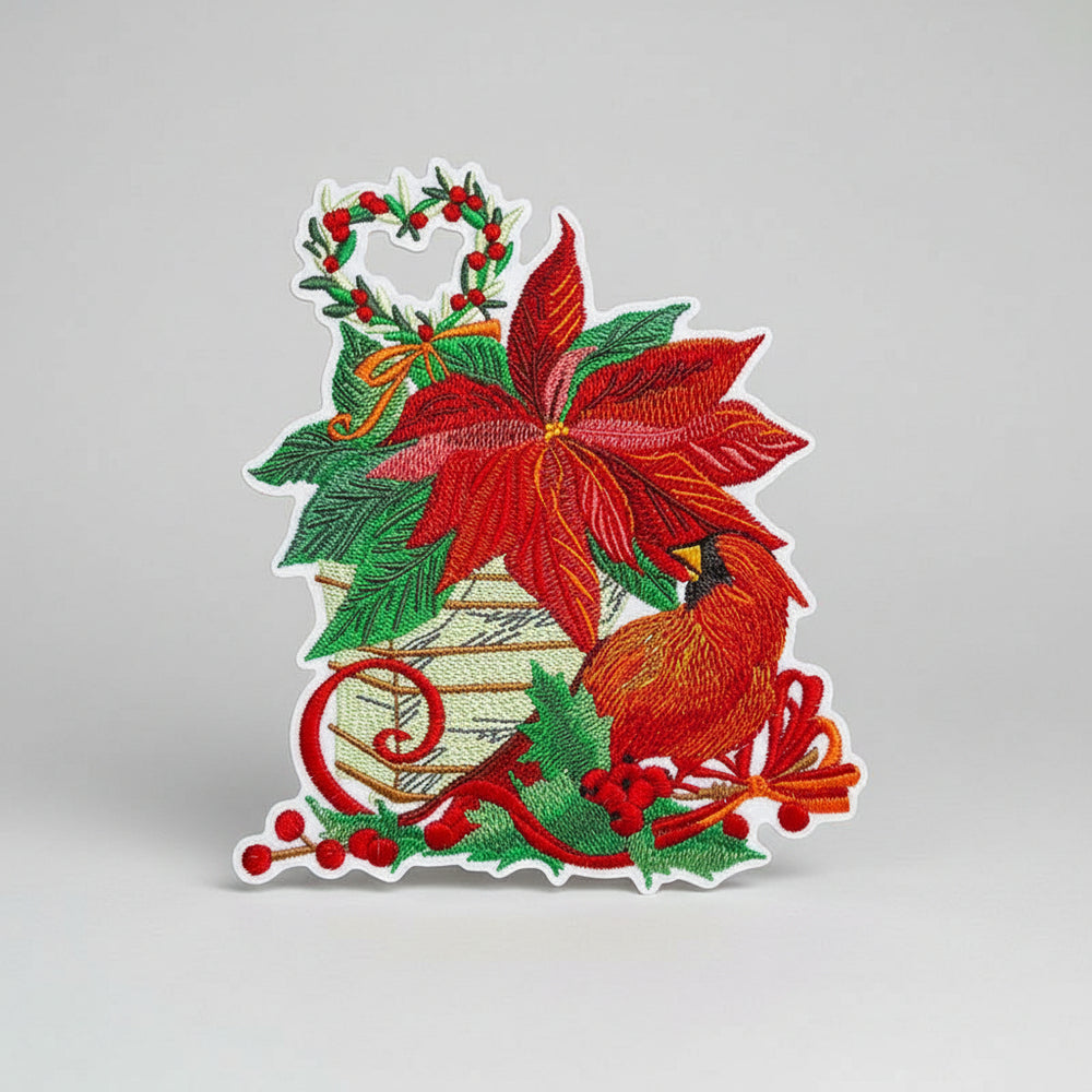 Close-up of embroidered poinsettia arrangement with a red cardinal, holly berries, pine leaves, and a heart-shaped wreath on white fabric.
