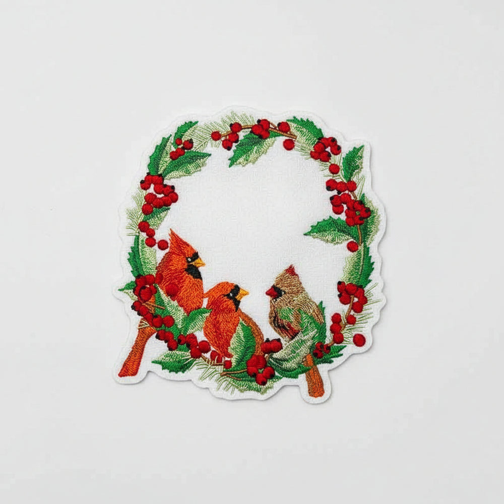 Embroidered design featuring two red cardinals among holly and berries on a white fabric.