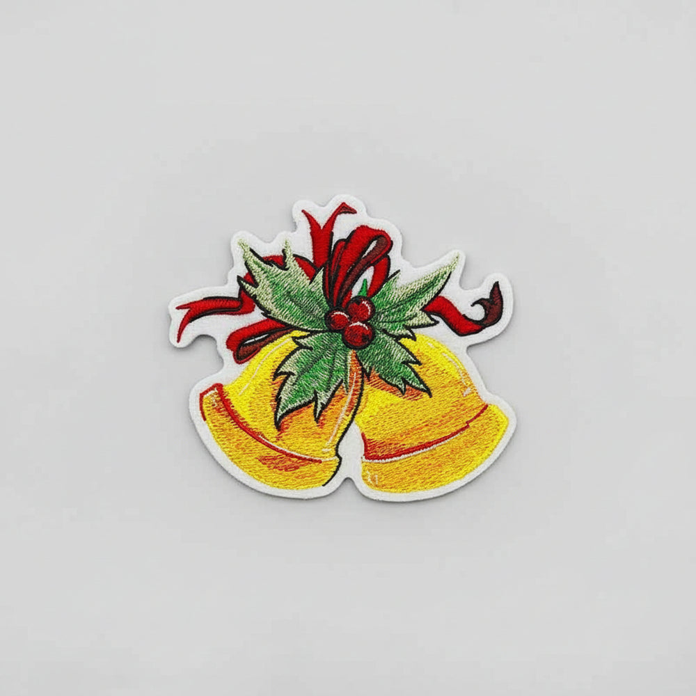 Close-up of embroidered golden Christmas bells adorned with green holly leaves, red berries, and a red ribbon on white fabric.