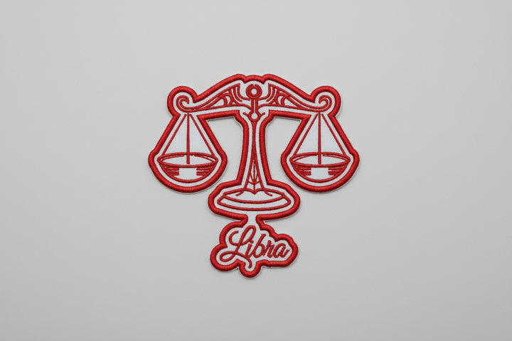 Libra zodiac scales embroidery patch featuring a balanced scales design in red outline stitching and the word “Libra” in script below, on a light background, original EmbroMania artwork.