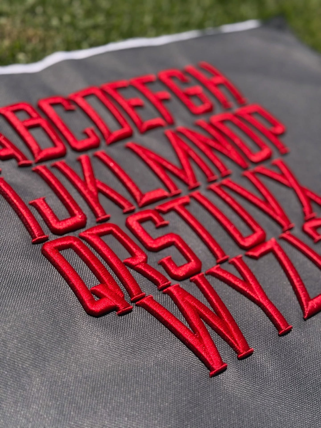  Close-up of red satin-stitch embroidered Metro Block alphabet letters (A–Z) arranged in rows on dark gray fabric, photographed outdoors on grass.