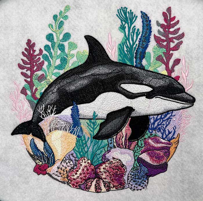 Embroidered design of an orca whale surrounded by colorful underwater plants on a light gray background.