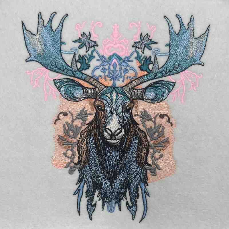 A stitched embroidery design of a moose head with ornate antlers and a floral pattern in blue, gray, and pink colors on a beige background.
