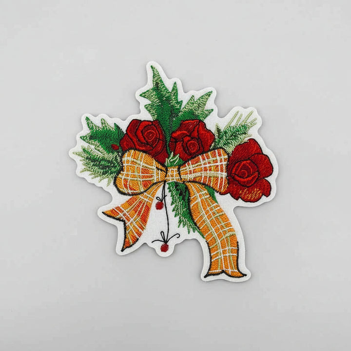 Embroidered design featuring red roses, green foliage, and an orange plaid bow on a light background.