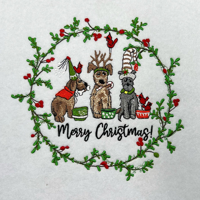 Close-up of embroidered Christmas wreath with three festive dogs and red cardinals, above the text “Merry Christmas!” on white fabric.