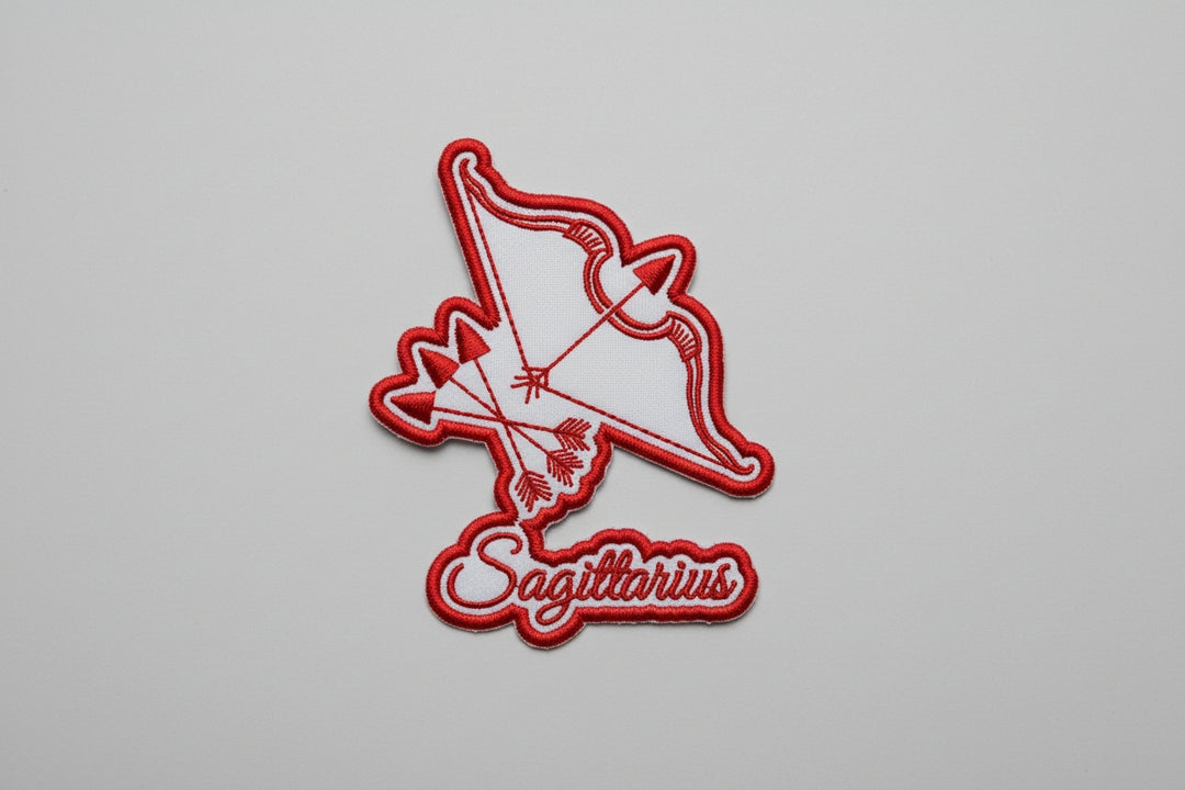 Sagittarius zodiac archer embroidery patch featuring a stylized bow and arrow design in red outline stitching and the word “Sagittarius” in script below, on a light background, original EmbroMania artwork.
