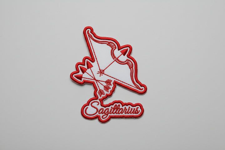 Sagittarius zodiac archer embroidery patch featuring a stylized bow and arrow design in red outline stitching and the word “Sagittarius” in script below, on a light background, original EmbroMania artwork.