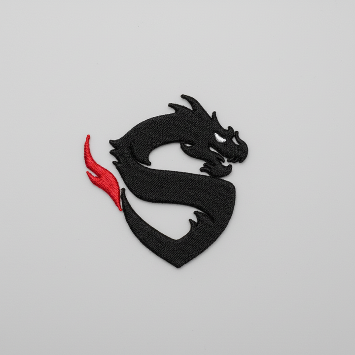 Minimal dragon embroidery patch featuring a black dragon silhouette with a sharp white eye and a red flame accent on the tail, stitched on a light background, original EmbroMania artwork.