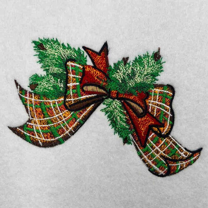 Close-up of embroidered tartan plaid bow with green pine sprigs on white fabric.