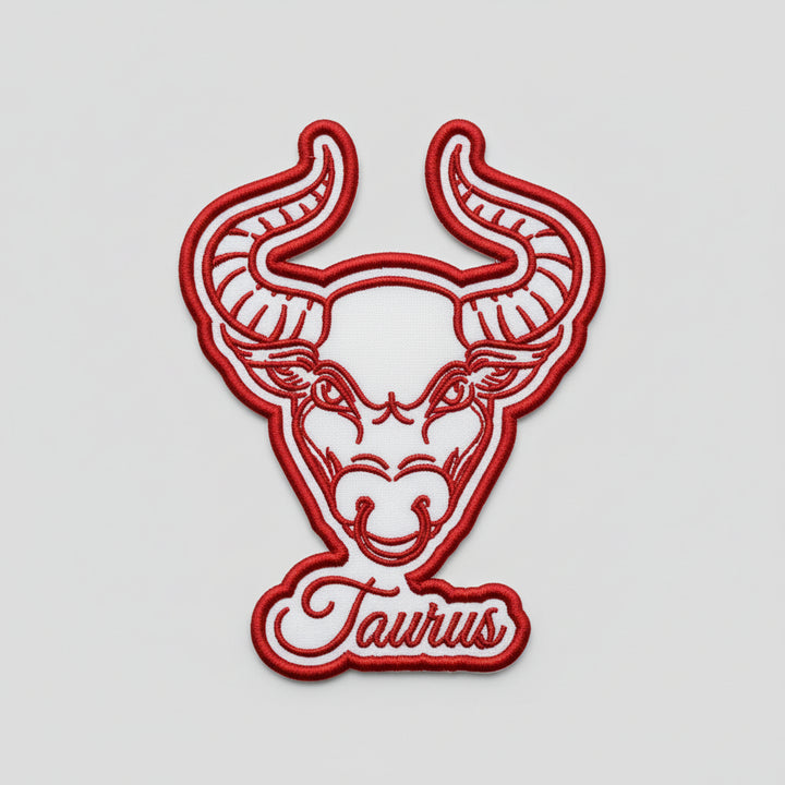 Taurus zodiac bull head embroidery patch featuring a bold bull face with curved horns in red outline stitching and the word “Taurus” in script below, on a light background, original EmbroMania artwork.