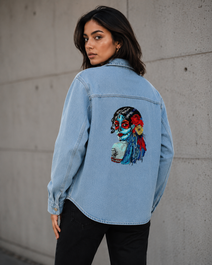 Woman wearing a light blue denim jacket with a colorful graphic on the back against a concrete wall.