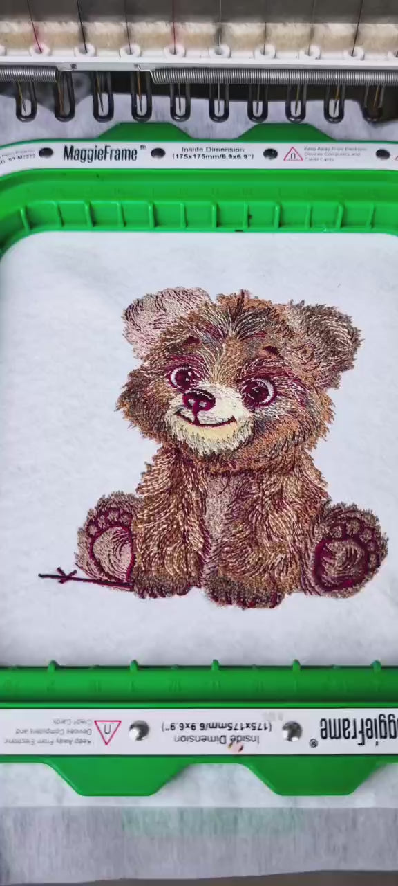 Vertical video showing a baby brown bear embroidery being stitched on a machine in a green hoop, with close-ups of the needlework and a final EmbroMania “Crafting Visions” logo screen.