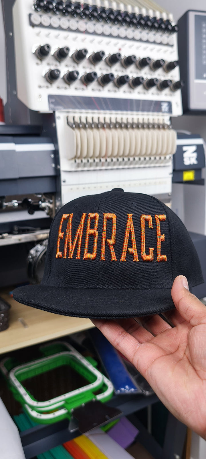 Close-up of a black cap featuring “EMBRACE” in Metro-style multicolor 3D puff embroidery, shown during stitching on an embroidery machine, ending with the Embromania logo and “Crafting Visions” text.